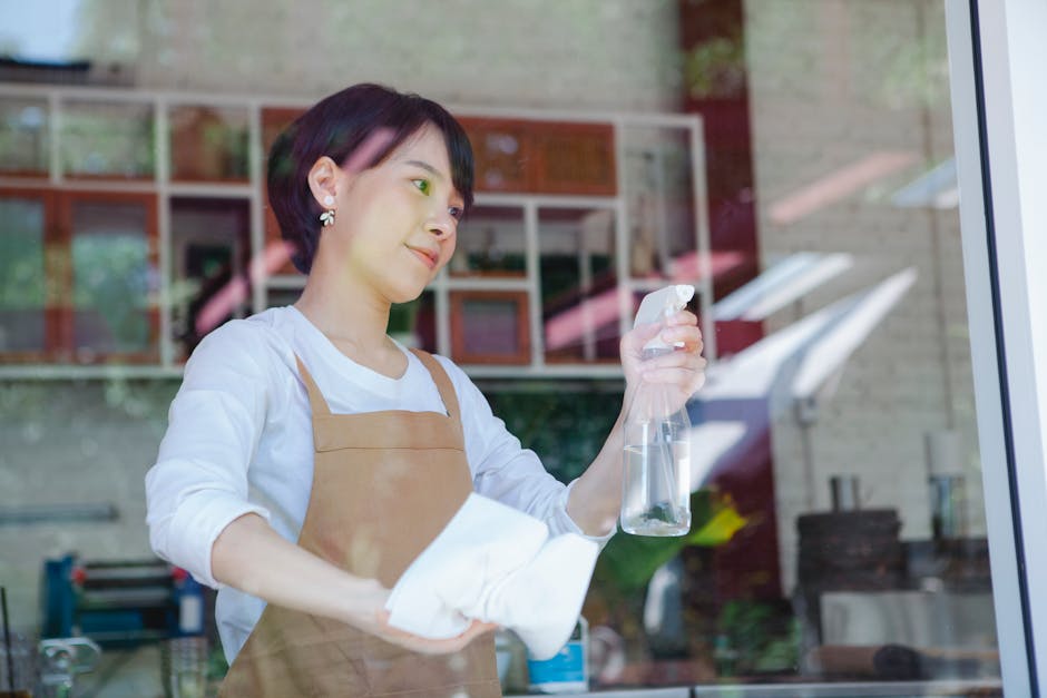 A woman with short dark hair, wearing a white t-shirt and brown apron, is engaged in surface cleaning inside a modern café or small restaurant. She is leaning forward, using a cloth to wipe a wooden 