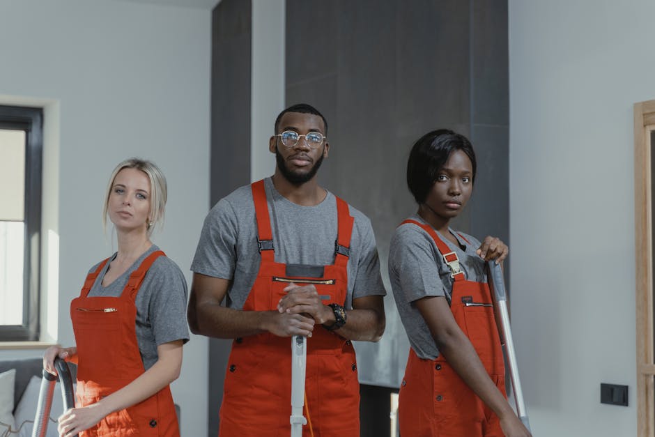 Three professional cleaners from Alperton Carpet Cleaners stand indoors in a modern, well-lit room with a grey wall and a window. They are wearing matching grey shirts and bright orange aprons, holdin