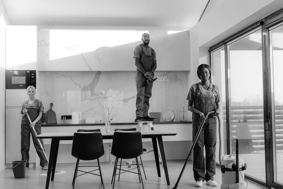 Three professional cleaners from Alperton Carpet Cleaners stand indoors in a modern, well-lit room with a grey wall and a window. They are wearing matching grey shirts and bright orange aprons, holdin